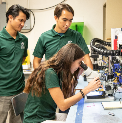 Three people in green shirts are in a lab. One person is using a microscope while the others observe. Equipment and wires can be seen on the table.